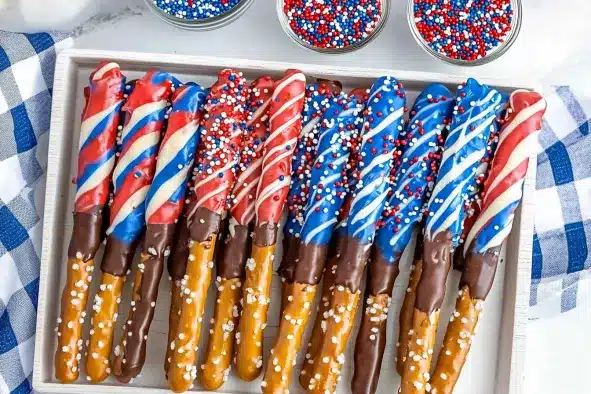 Patriotic chocolate-covered pretzel rods arranged in white wooden tray with red, white, and blue candy coatings and sprinkle decorations, three small glass bowls of sprinkles and blue checkered towel visible