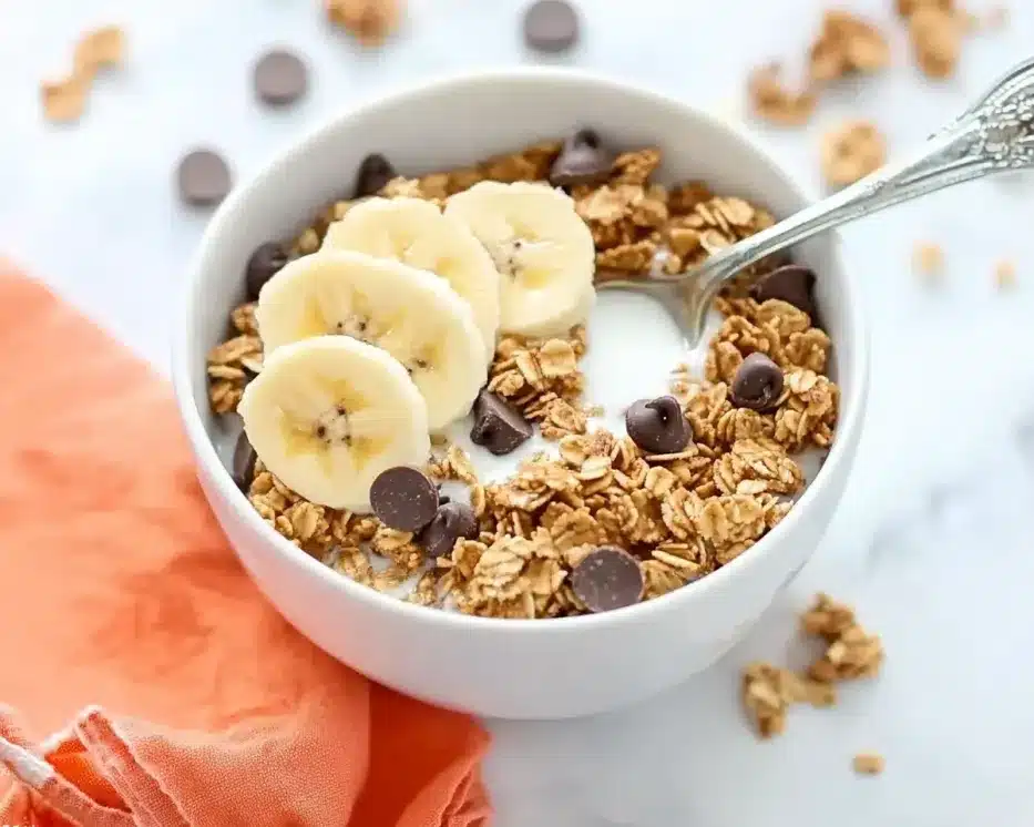 White ceramic bowl filled with peanut butter granola, topped with fresh banana slices and chocolate chips, milk added, spoon ready for eating