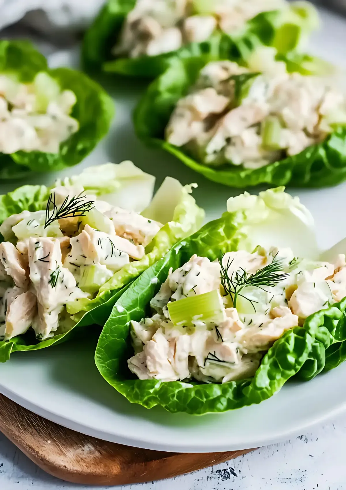 Two keto chicken salad lettuce wraps on white plate with wooden trivet, featuring chunky chicken salad and fresh dill garnish