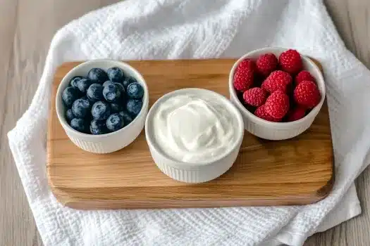 Overhead view of vegan frozen yogurt cups preparation on a wooden board with teal bowls of fresh blueberries and raspberries, white paper cupcake liners filled with yogurt, and a bowl of vegan yogurt on marble countertop.