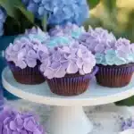 Chocolate cupcakes with purple and blue hydrangea-style frosting on a white cake stand with real hydrangeas in background.