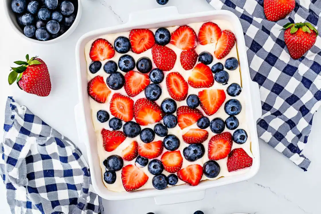 Overhead view of No Bake Berry Icebox Cake in white dish with halved strawberries and whole blueberries arranged on cream topping, blue gingham napkin, white marble background