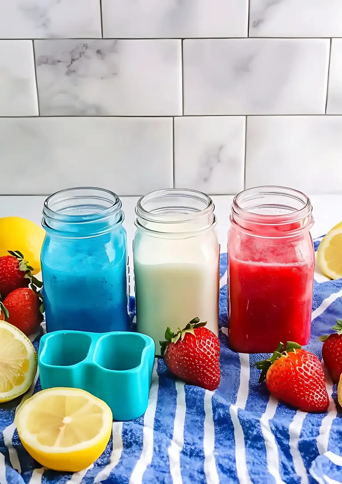 Three mason jars containing red strawberry puree, white coconut milk, and blue raspberry mixture with fresh strawberries and lemons on a blue striped cloth