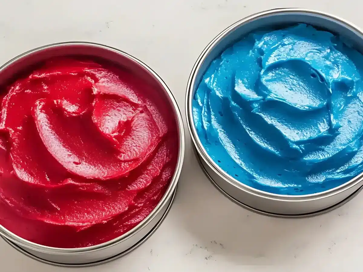 Two round cake pans containing vibrant red and electric blue cake batter on a white marble surface