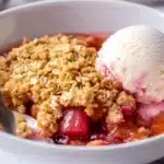 Close-up of rhubarb crisp serving in white bowl with vanilla ice cream scoop, golden crumble topping and vibrant red rhubarb filling visible with spoon