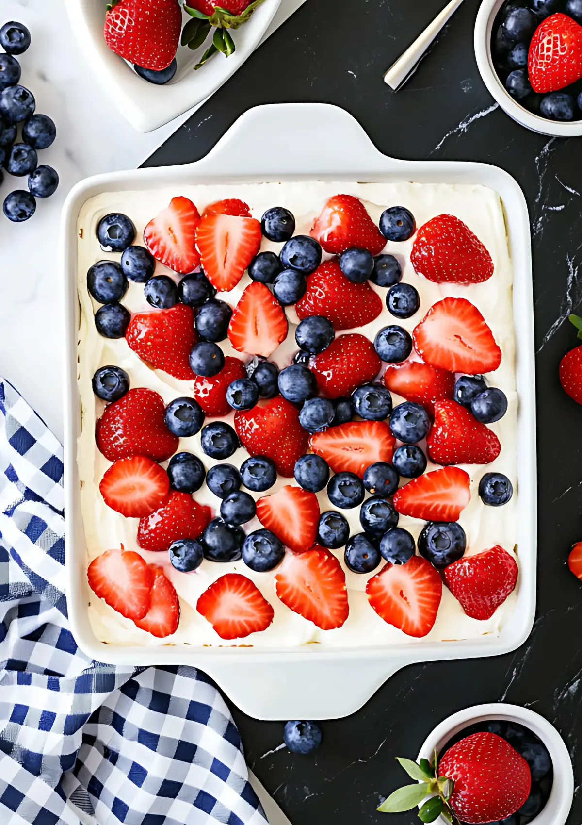 Overhead view of No Bake Berry Icebox Cake in white dish on black marble countertop, decorated with fresh strawberries and blueberries, blue gingham napkin
