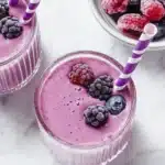 Top view of a berry smoothie in a clear glass topped with frozen blackberries and blueberries, next to scattered berries on a white surface.