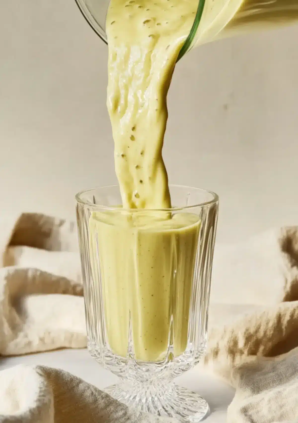 Pouring thick pistachio smoothie from blender into a crystal footed glass with linen backdrop.
