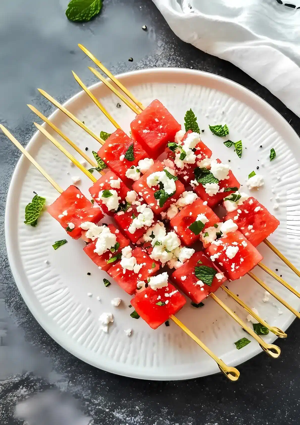Golden skewers loaded with watermelon cubes, mint, and feta arranged on a white textured plate, styled for elegant summer entertaining