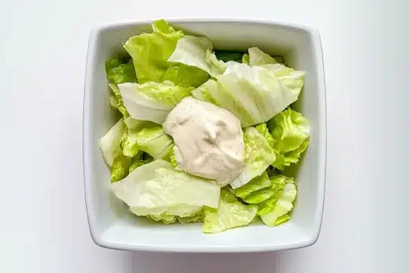 Fresh iceberg lettuce leaves in white square bowl with dollop of creamy white dressing in center, ready for mixing