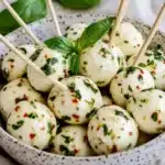 Marinated mozzarella balls with toothpicks in speckled ceramic bowl, garnished with fresh basil leaves and red pepper flakes
