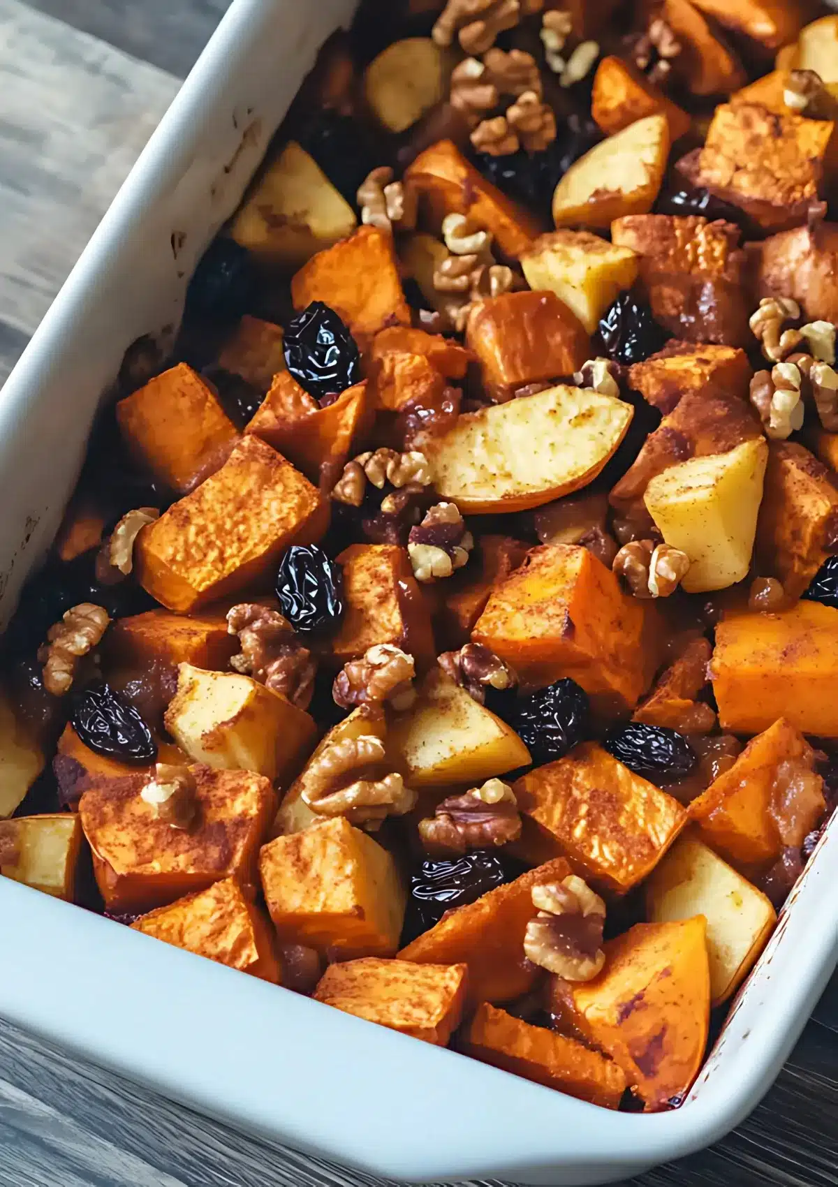 Angled view of apple sweet potato bake in white rectangular baking dish showing perfectly roasted vegetables, nuts, and dried fruit with caramelized edges