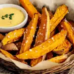 Sweet potato fries arranged in a wicker basket lined with parchment paper, seasoned with salt and served alongside creamy herb aioli