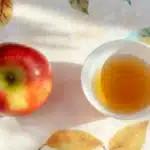 A fresh red apple placed next to a white bowl filled with golden apple cider vinegar on a light floral-patterned tablecloth with soft morning sunlight.