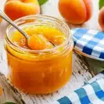 Open jar of chunky apricot jam with spoon, fresh whole and halved apricots, blue checkered cloth on rustic wooden surface