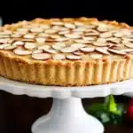 Golden frangipane tart topped with sliced almonds, displayed on a scalloped white cake stand with raspberries and mint in the background.