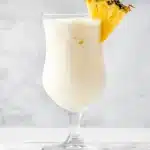 A Piña Colada cocktail in a hurricane glass with crushed ice, garnished with a fresh pineapple wedge, set against a light marble background.