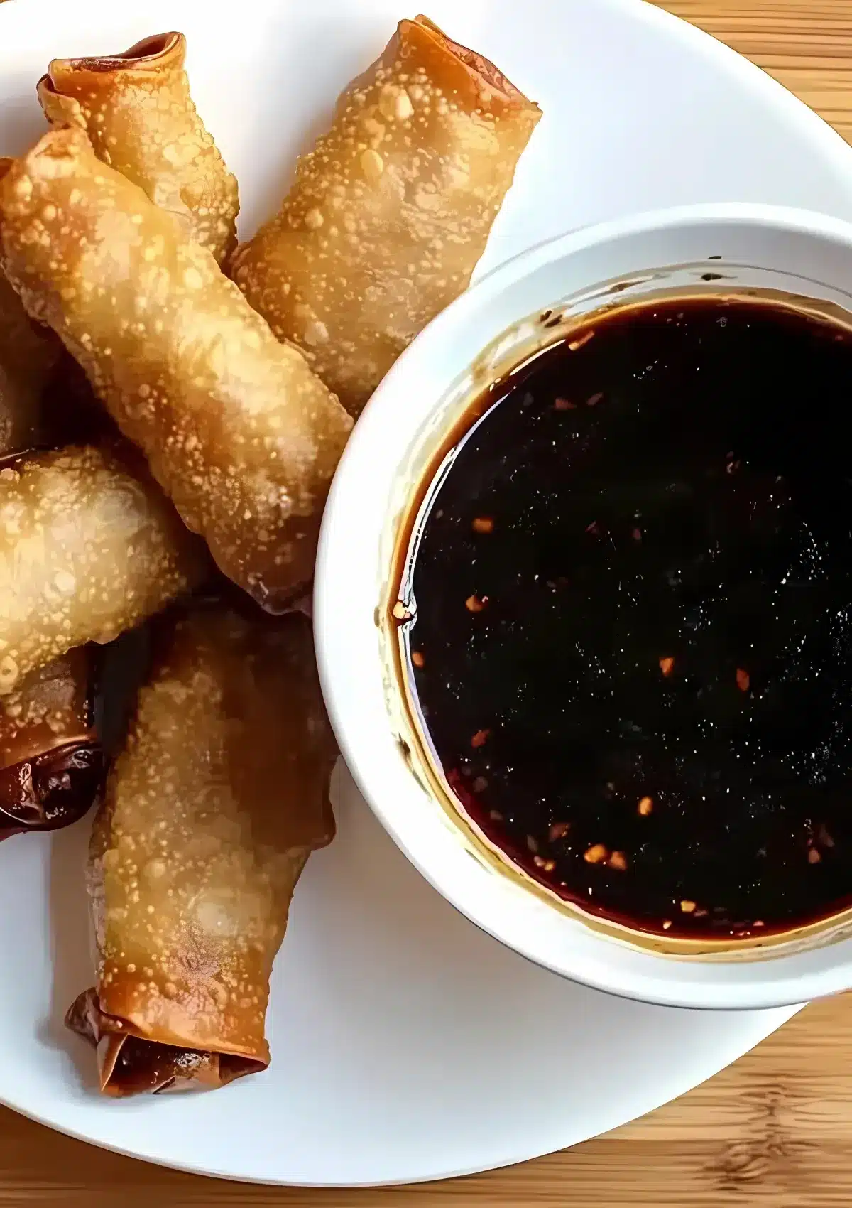 Close-up of crunchy egg rolls next to a bowl of thick Mongolian sauce, served on a round white plate over a wooden table

