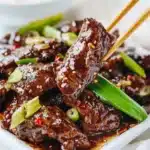 Chopsticks holding a piece of glossy Mongolian beef garnished with scallions and sesame seeds on a white plate.
