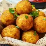 Golden brown cheesy mashed potato balls served in a rustic paper-lined basket, garnished with parsley, perfect for dipping in ketchup.