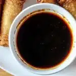 Overhead view of golden crispy egg rolls arranged around a white bowl filled with glossy Mongolian sauce on a white plate