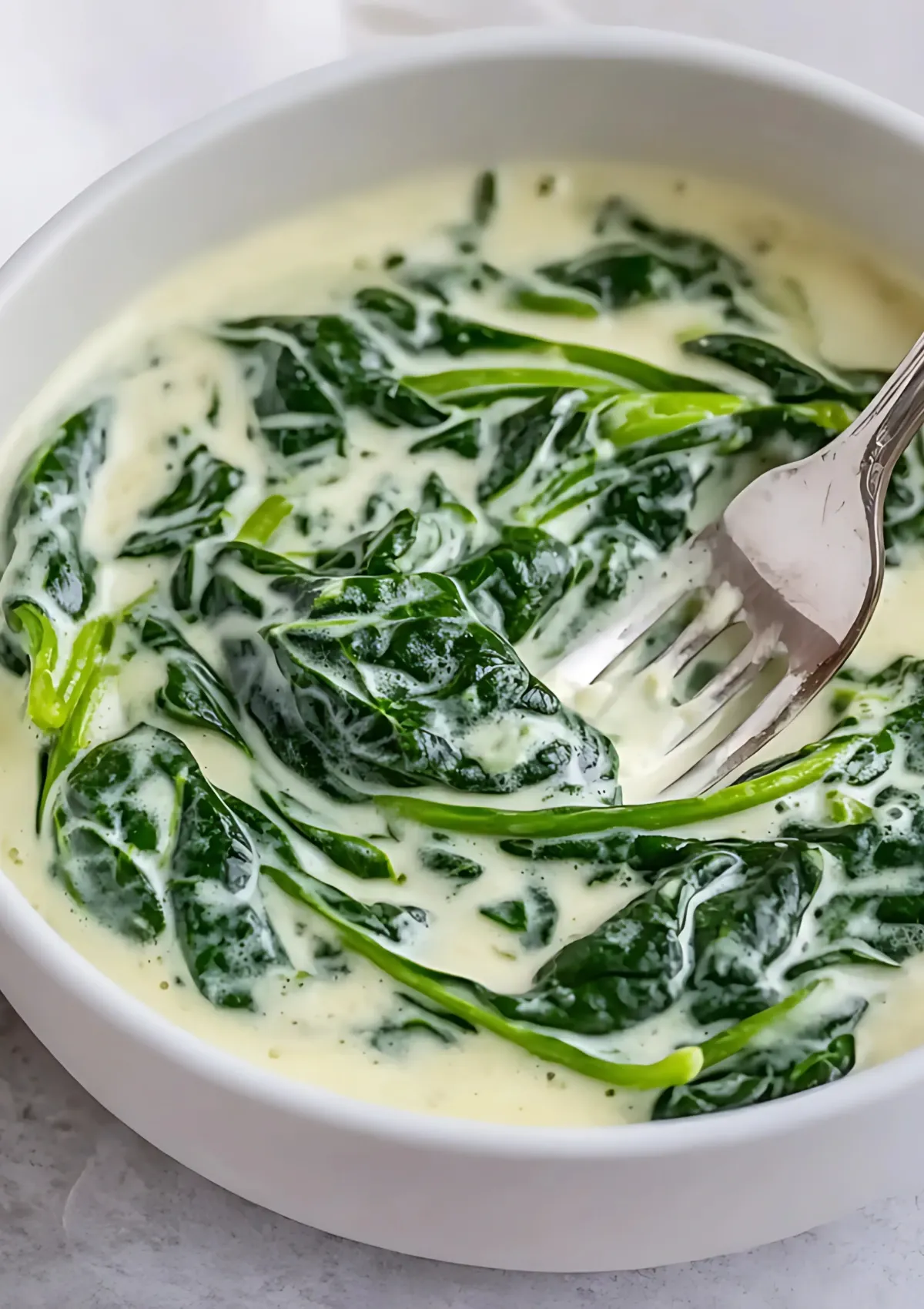 Serving dairy free creamed spinach with fork from white bowl, showing creamy plant-based sauce
