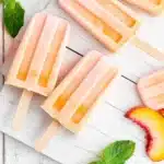 Flat lay of peach popsicles on white wooden boards with fresh peach slices and mint leaves