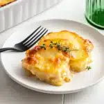 A portion of golden scalloped potatoes served on a white plate with a black fork, white baking dish in background, showing the creamy layers and browned top