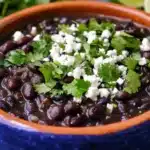 Blue ceramic bowl containing Mexican black beans seasoned with cumin and topped with fresh cilantro and crumbled white cheese