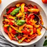 Overhead shot of pepperonata recipe in ceramic bowl surrounded by fresh cherry tomatoes, basil leaves, and capers