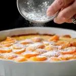 Hand dusting powdered sugar over finished apricot flaugnarde dessert with golden custard and caramelized apricots