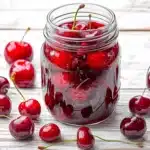 Fresh tipsy cherries in mason jar with scattered cherries on white wooden surface