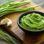 Garlic scape pesto served in small black bowl on wooden board with fresh garlic scapes and garlic cloves as garnish
