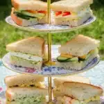 A three-tier cake stand outdoors, holding fresh vegetarian tea sandwiches filled with cucumber, cheese, eggs, and peppers, displayed on a floral picnic blanket under sunlight.