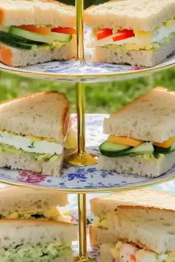 A three-tier cake stand outdoors, holding fresh vegetarian tea sandwiches filled with cucumber, cheese, eggs, and peppers, displayed on a floral picnic blanket under sunlight.