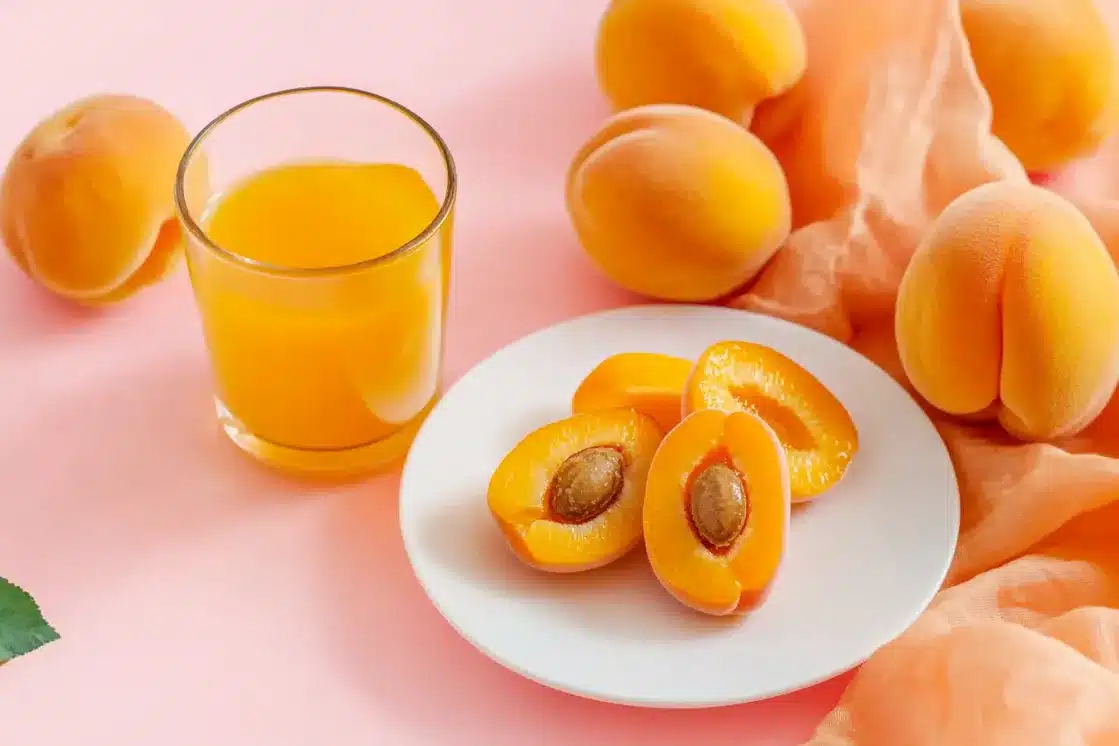 Fresh ripe apricots whole and halved on white plate, glass of apricot juice, pink background with scattered apricots