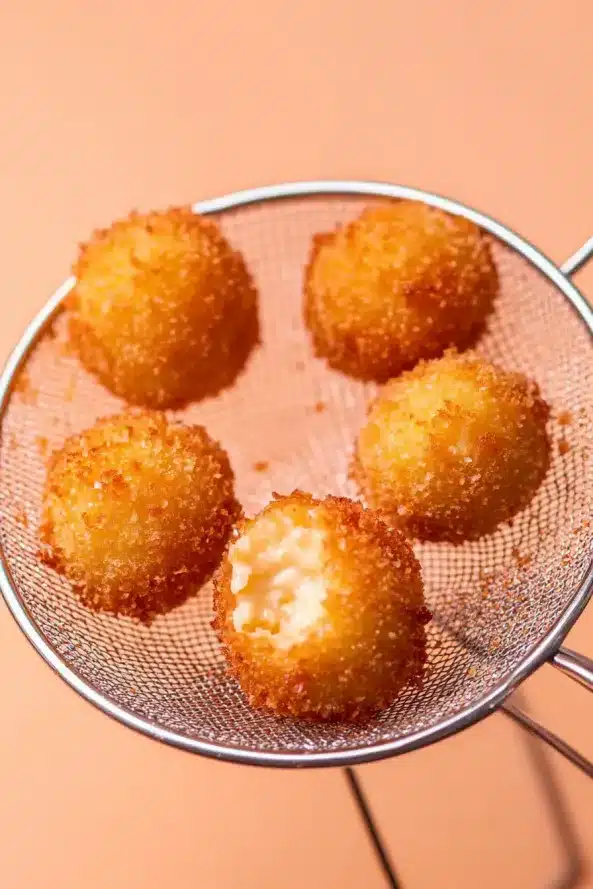 Five crispy golden potato balls, one partially bitten, resting in a frying strainer over a peach-colored surface.