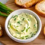 Garlic scapes butter spread on bread slices with bowl of compound butter and fresh garlic scapes on cutting board