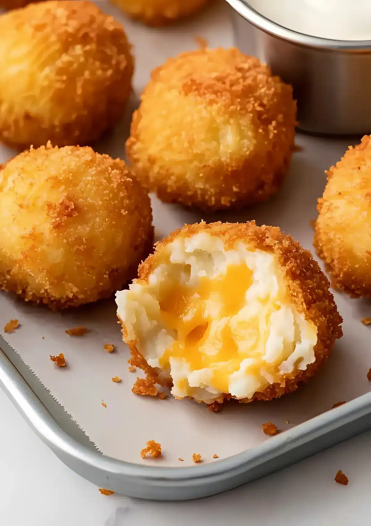Fried potato balls on a tray with one cut open revealing melted cheddar cheese, served alongside a metal dipping sauce cup.