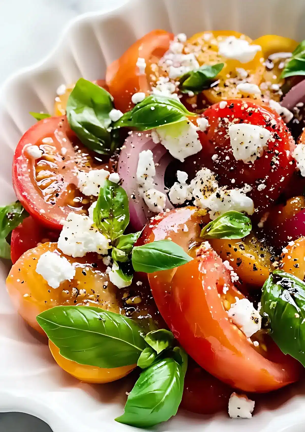 Close-up view of colorful heirloom tomato salad on white plate showing detailed texture of mixed tomato varieties, white feta cheese, fresh basil leaves, and black pepper seasoning