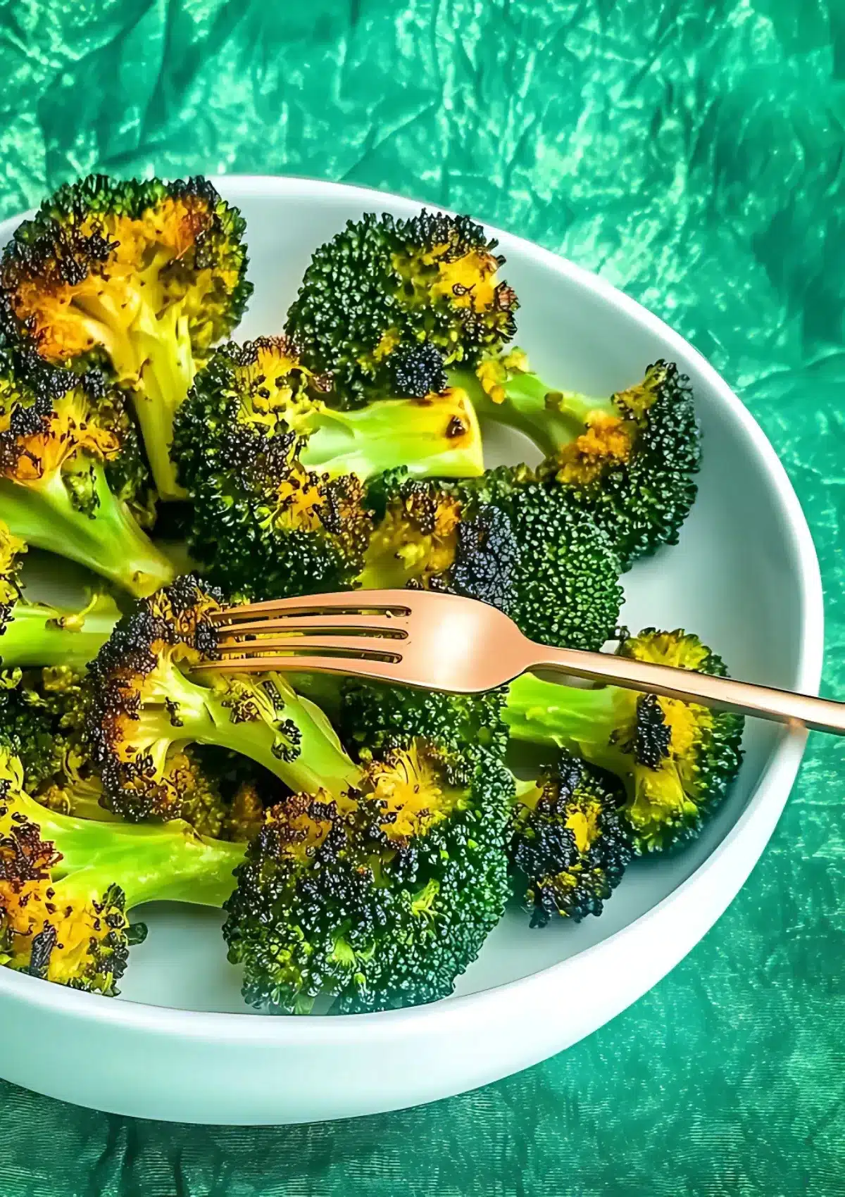 Golden grilled broccoli in white bowl with rose gold fork on vibrant green background