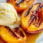 A close-up of perfectly grilled peach halves with caramelized marks, served with a scoop of vanilla ice cream and drizzled with golden syrup on a white plate.