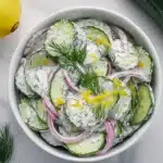 Creamy cucumber salad in white bowl with fresh dill garnish, lemon and cucumber in background