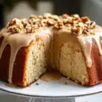 Maple cream cake on white plate with maple syrup glaze and chopped walnuts, cut to show fluffy cake interior