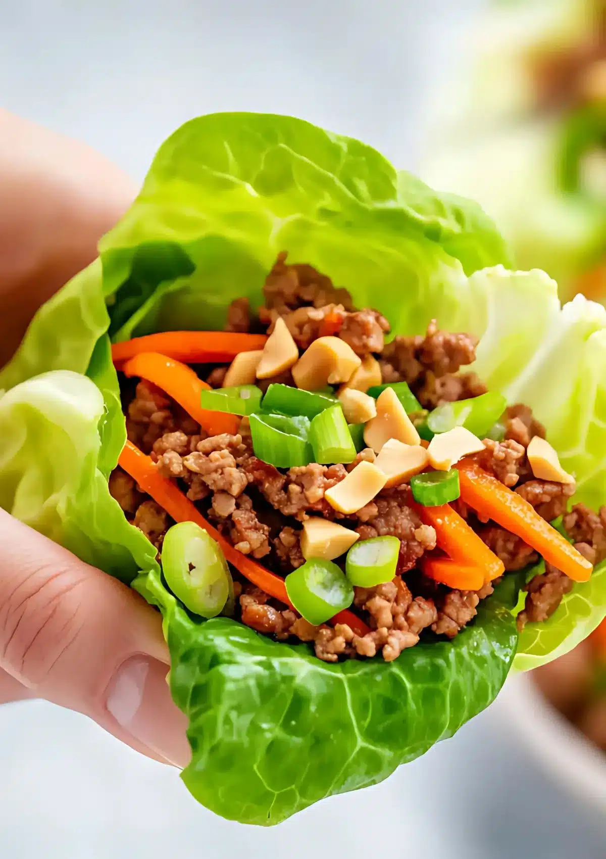 Close-up of hands holding a single orange beef lettuce wrap showing the colorful filling with ground beef, orange carrots, green onions,