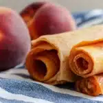 Golden peach leather rolls on a blue striped kitchen towel with fresh peaches in the background