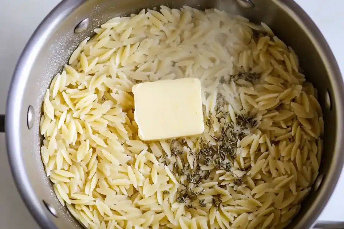Cooked orzo pasta in stainless steel pot with melted butter and Italian seasoning being stirred together