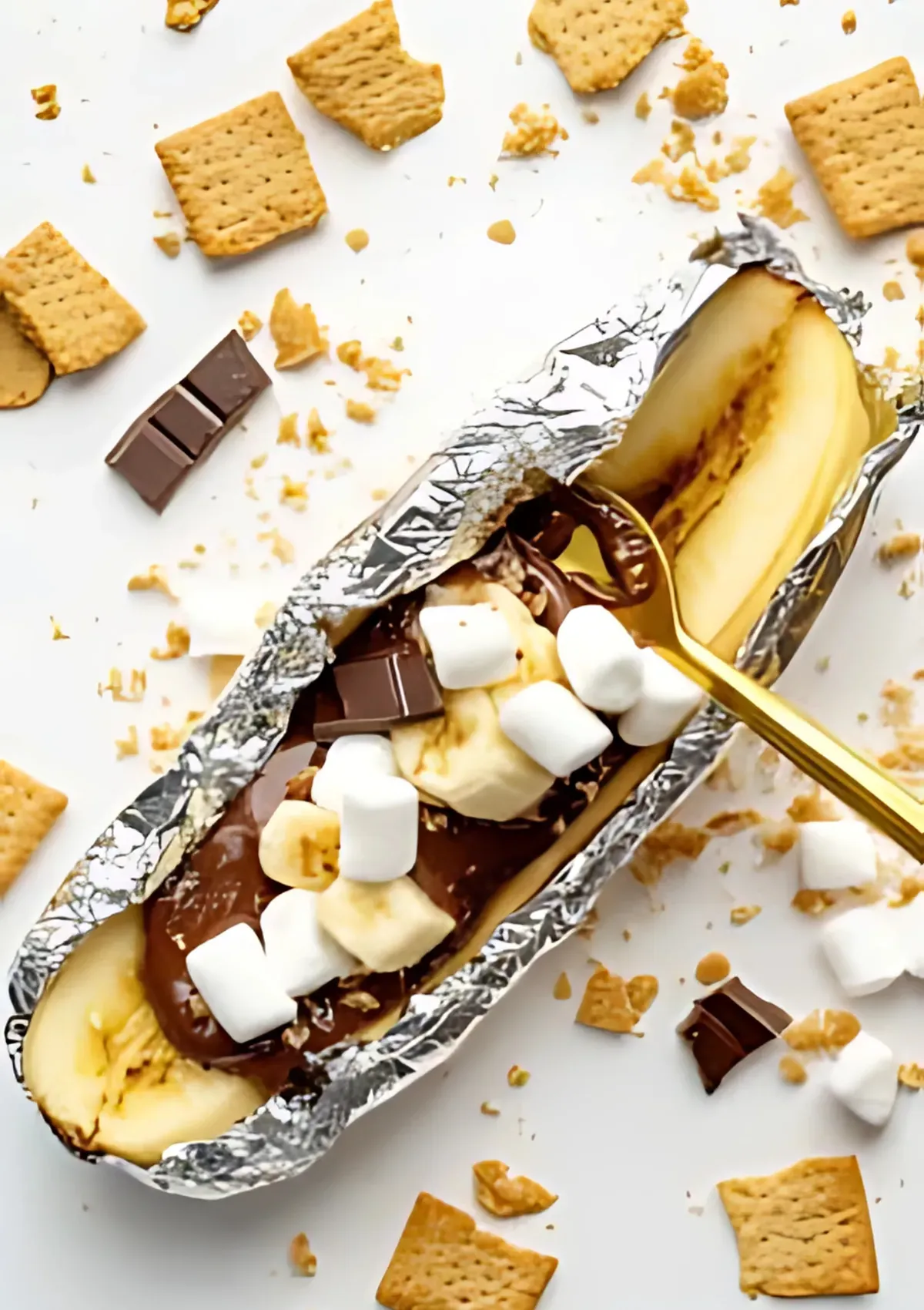Overhead view of a banana boat topped with mini marshmallows and chocolate chunks, ready to be baked in foil with golden spoon on a white background.

