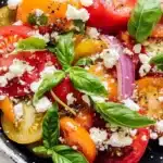 Mediterranean heirloom tomato salad in speckled ceramic bowl with sliced multicolor tomatoes, white feta cheese, fresh basil, red onions, and herb seasoning