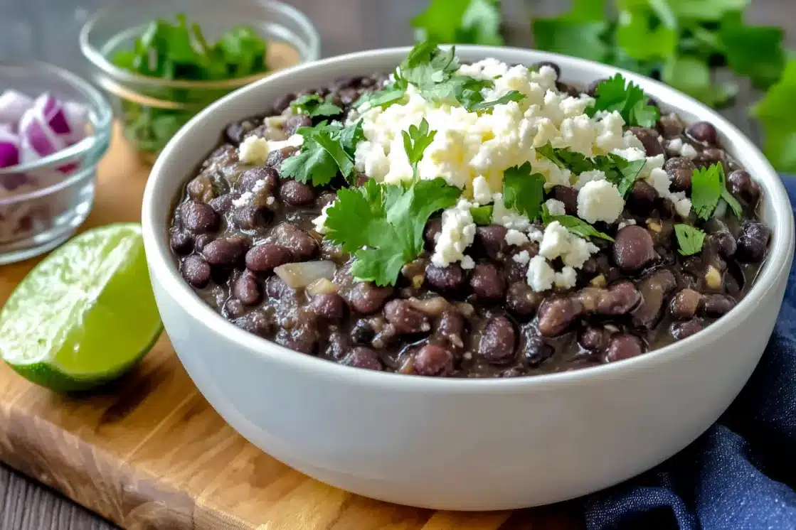 White bowl of Mexican black beans garnished with cilantro and cheese, served with lime wedges and fresh herbs on wooden board